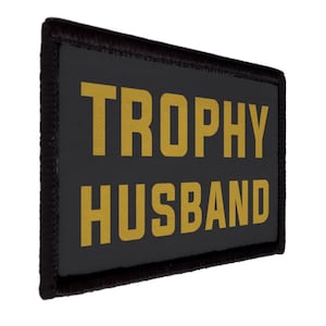 Peut inclure: Un écusson rectangulaire noir avec les mots "TROPHY HUSBAND" en lettres dorées. L'écusson a une bordure noire et est posé sur un fond blanc.