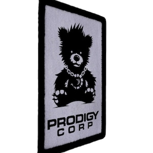 Könnte beinhalten: Ein rechteckiger Aufnäher mit schwarzem Rand und hellgrauem Hintergrund. Der Aufnäher zeigt eine schwarze Silhouette eines Teddybären mit einer Halskette mit einem Mondsichel-Anhänger. Der Text "PRODIGY CORP" ist in Weiß gedruckt.