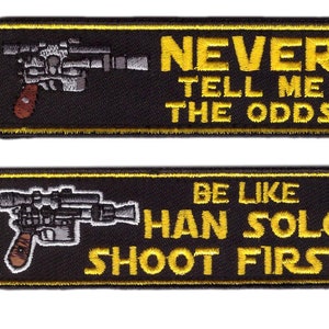 Könnte beinhalten: Zwei schwarze rechteckige Aufnäher mit gelbem Text und einem silbernen und braunen Blaster-Design. Der obere Aufnäher lautet: "NEVER TELL ME THE ODDS". Der untere Aufnäher lautet: "BE LIKE HAN SOLO SHOOT FIRST."