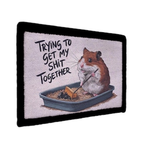 Peut inclure: Écusson rectangulaire à bordure noire. Il représente un hamster brun et blanc dans une litière, tenant un bâton. Le texte se lit "Trying to get my shit together."