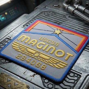 May include: A rectangular embroidered patch featuring a gray background with a blue border. The text reads USCSS MAGINOT 294068 in gold thread. A gold winged emblem sits below the text, and a yellow star with radiating lines sits above.