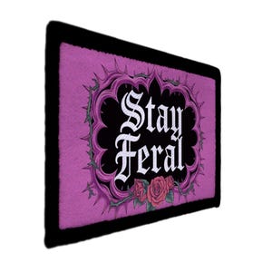 Pode incluir: Um tapete retangular roxo com as palavras "Stay Feral" em fonte gótica. O texto está dentro de uma moldura preta ornamentada com espinhos e rosas. O tapete tem uma borda preta.