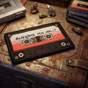 Könnte beinhalten: Gestickter Aufnäher in Form eines Kassettenbands mit dem Text "AWESOME MIX VOL. 1" in Blau. Das Banddesign hat rote und weiße Streifen und einen schwarzen Rand. Weitere Kassettenbänder und Kristalle befinden sich im Hintergrund.