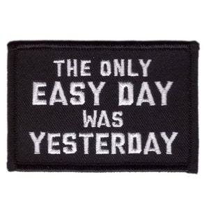May include: Black rectangular patch with white embroidered text that reads "THE ONLY EASY DAY WAS YESTERDAY". The patch has a black border and is likely designed to be sewn or ironed onto clothing or accessories.
