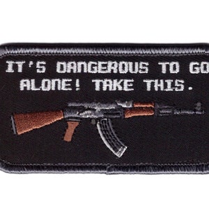 May include: Black rectangular patch with the text "IT'S DANGEROUS TO GO ALONE! TAKE THIS." A stylized image of an AK-47 rifle is below the text. The rifle has brown and gray details. The patch has a black border.