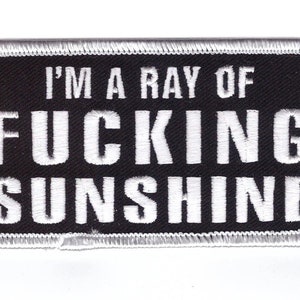 May include: Black rectangular patch with white embroidered text that reads "I'M A RAY OF FUCKING SUNSHINE". The patch has a white stitched border.