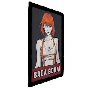 Op de afbeelding: Een rechthoekige patch met een zwarte rand met een gestileerde illustratie van een vrouw met rood haar en oranje bretels. De tekst "BADA BOOM" wordt in witte letters op een rode banner weergegeven.