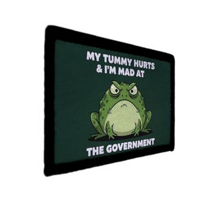 Könnte beinhalten: Ein dunkelgrüner Aufnäher mit schwarzem Rand. Der Aufnäher zeigt einen Cartoon-Frosch mit wütendem Gesichtsausdruck. Der Text auf dem Aufnäher lautet "MY TUMMY HURTS & I'M MAD AT THE GOVERNMENT."