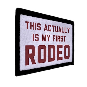 Könnte beinhalten: Rechteckiger Aufnäher mit weißem Hintergrund und schwarzem Rand. Der Text "THIS ACTUALLY IS MY FIRST RODEO" ist in weinroten, fetten Buchstaben gedruckt. Der Aufnäher ist wahrscheinlich für Kleidung oder Accessoires.