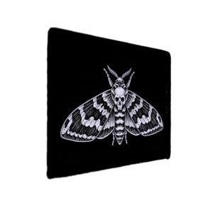 May include: Black mousepad featuring a detailed white illustration of a moth with a skull-like head and patterned wings. The moth design is centered on the black surface, creating a striking contrast. The mousepad is rectangular.