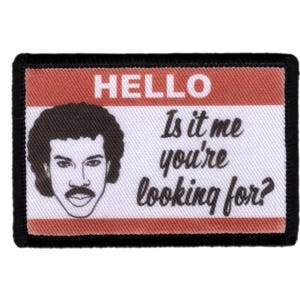 Peut inclure: Écusson rectangulaire à bordure noire, avec un portrait et le texte "HELLO Is it me you're looking for?" dans un design de badge nominatif. La partie supérieure est rouge et le bas est blanc.