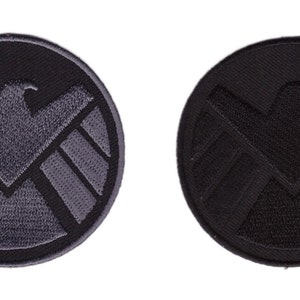 Pode incluir: Dois patches redondos bordados. Um patch é cinza escuro com uma borda preta e apresenta um emblema de águia estilizado dentro de uma forma de escudo. O outro patch é preto com um emblema preto.