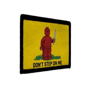 May include: A yellow patch with a black border features a red Lego figure holding a sword, standing on green grass. The text "DON'T STEP ON ME" is printed in black below the figure. The patch is square.