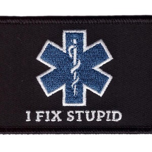 May include: Black rectangular patch with a blue and white medical symbol, the Star of Life, embroidered in the center. The text "I FIX STUPID" is embroidered in white below the symbol. The patch has a black border.