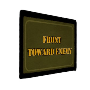 Peut inclure: Un écusson rectangulaire vert olive avec une bordure noire épaisse. Le texte FRONT TOWARD ENEMY est imprimé au centre dans une police jaune grasse de style pochoir. L'écusson a une surface texturée et est vu sous un angle latéral.