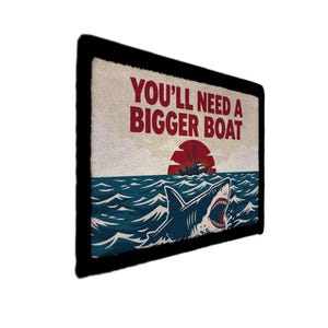 Op de afbeelding: Rechthoekige patch met een zwarte rand met een haai in de oceaan met de tekst "YOU'LL NEED A BIGGER BOAT". Het ontwerp bevat een rode zonsondergang en een boot aan de horizon. De haai is blauw en wit.
