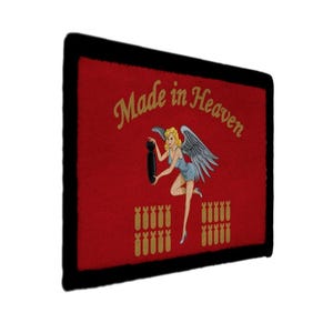 May include: A rectangular patch with a red background and a black border. The patch features the text "Made in Heaven" in gold script, along with an illustration of a blonde woman with wings holding a bomb. Below are rows of gold bombs.
