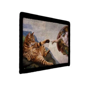 May include: A rectangular wall art print featuring a parody of The Creation of Adam, where a ginger tabby cat replaces Adam, reaching out its paw toward a figure on the right. The artwork is framed by a thick black border on all four sides.