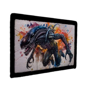 Op de afbeelding: Een rechthoekige grafiek met zwarte rand met een illustratie in aquarel van een Alien-wezen. Het wezen is afgebeeld in tinten zwart, blauw en grijs, met een gedetailleerde skeletstructuur en scherpe klauwen. De achtergrond heeft kleurrijke verfspatten.