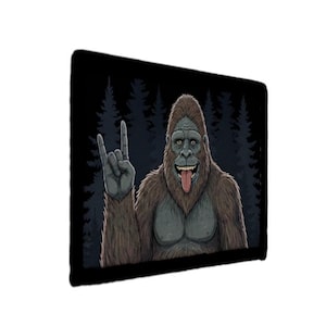 May include: A black mousepad with a cartoon illustration of a Bigfoot giving the rock and roll hand gesture. The Bigfoot has brown fur, a gray face, and a red tongue. A forest silhouette is in the background.