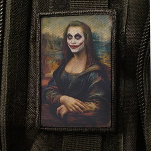 Pode incluir: Um patch retangular com uma paródia da Mona Lisa com maquiagem do Coringa. O patch está em um fundo preto. O rosto da Mona Lisa é pintado com maquiagem branca, vermelha e verde, imitando a aparência do Coringa.