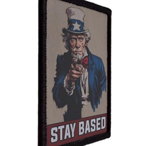 May include: A rectangular patch with an illustration of Uncle Sam pointing, wearing a blue suit and a hat with a star. The text "STAY BASED" is displayed in white on a red banner. The patch has a black border.