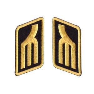 May include: Two embroidered patches with a black background and a gold-colored design. The design features a stylized shape with three vertical lines. The patches have a gold border and are set against a white background.