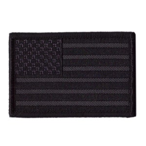 Op de afbeelding: Een rechthoekige zwarte patch met een ingetogen ontwerp van de Amerikaanse vlag. De sterren en strepen zijn in tinten zwart weergegeven, wat een monochroom effect creëert. De patch heeft een zwarte rand en is waarschijnlijk gemaakt van stof.