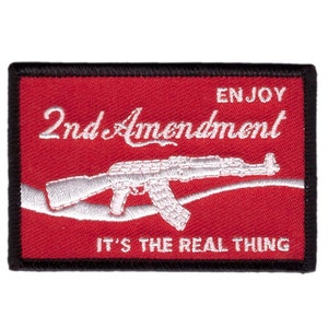 Op de afbeelding: Een rechthoekige patch met een rode achtergrond en een zwarte rand. De patch toont de witte tekst "ENJOY 2nd Amendment IT'S THE REAL THING" en een wit geborduurd beeld van een geweer.