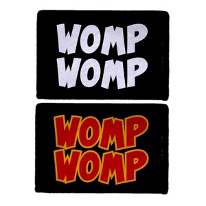 May include: Two black rectangular patches with the word "WOMP" in white and red with yellow outline. The text is in a bold, comic book style font. The patches are displayed against a white background.