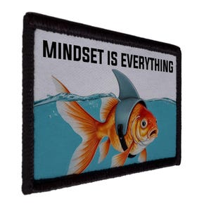 May include: A rectangular patch with a black border features a goldfish wearing a shark fin hat, submerged in blue water. The text "MINDSET IS EVERYTHING" is printed in bold, black letters above the fish. The patch has a white background.