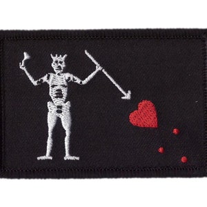 Op de afbeelding: Zwart rechthoekig embleem met een wit geborduurd skeletfiguur met een speer en een beker. Een rood hart en drie rode stippen bevinden zich rechts van het skelet. Het embleem heeft een zwarte rand.