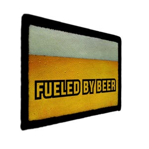 May include: A rectangular patch with a black border, featuring a beer-themed design. The top portion is light beige, transitioning to a golden-yellow section below. Bold, black text reads "FUELED BY BEER".