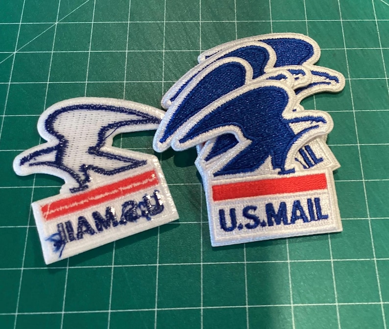 Puede incluir: Parches bordados con el logotipo de U.S. Mail, un dise&ntilde;o de &aacute;guila azul, detalles en rojo y blanco, y el texto "U.S. MAIL". Los parches est&aacute;n sobre una superficie de cuadr&iacute;cula verde.