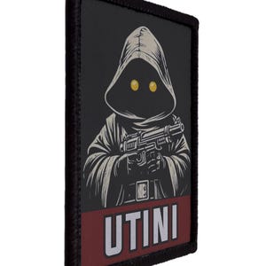 Pode incluir: Um patch retangular preto com uma figura encapuzada com olhos amarelos, segurando um blaster. A figura está em uma túnica bege. O patch tem uma faixa vermelha na parte inferior com a palavra "UTINI" em branco.