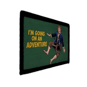 Op de afbeelding: Een groene patch met de tekst "I'M GOING ON AN ADVENTURE" in het geel. De patch toont een cartoon illustratie van een personage in een blauwe jas en bruine broek, halverwege een sprong. De patch heeft een zwarte rand.