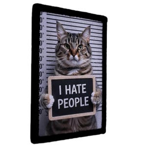 May include: A tabby cat with green eyes holding a sign that reads "I HATE PEOPLE". The cat is positioned in front of a mugshot background with black and white lines. The sign has white text.