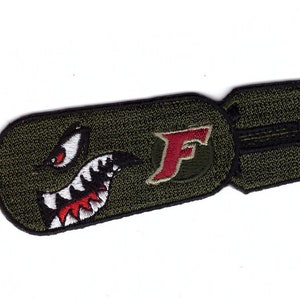 May include: Olive green embroidered bomb-shaped patch with a shark mouth design and the letter "F". The patch has black, white, and red detailing. It is likely designed to be sewn onto clothing or bags.