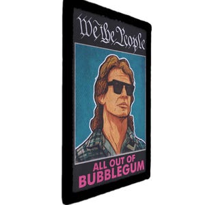 Op de afbeelding: Rechthoekige patch met een zwarte rand. Het toont een portret van een man met een zonnebril, met de tekst "We the People" erboven en "All Out of Bubblegum" eronder. De achtergrond is blauw.