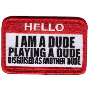 Pode incluir: Um patch retangular com borda vermelha e fundo branco. O patch diz "HELLO I AM A DUDE PLAYING A DUDE DISGUISED AS ANOTHER DUDE" em texto preto. O patch é provavelmente para passar a ferro ou costurar.