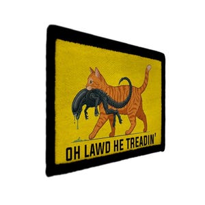 以下が含まれることがあります： 黒い縁取りと黄色の背景の長方形のパッチ。オレンジ色のトラ猫が歩いており、口に黒いエイリアンを運んでいます。「OH LAWD HE TREADIN'」というテキストが下にあります。