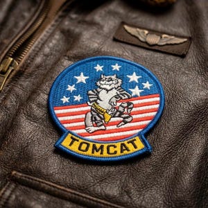 May include: Embroidered patch featuring a cartoon cat in a fighter pilot uniform, with the word "TOMCAT" in yellow. The patch has a blue background with white stars and red and white stripes. The patch is on a brown leather jacket.
