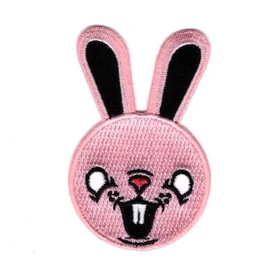Könnte beinhalten: Ein rosa bestickter Aufnäher in Form eines Hasengesichts. Der Hase hat schwarze Ohren, weiße Augen, eine rote Nase und einen schwarzen Mund mit zwei weißen Zähnen. Der Aufnäher ist kreisförmig und hat einen gestickten Rand.