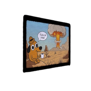 Op de afbeelding: Een rechthoekige, zwart omlijste print met een cartoonhond in een woestijnlandschap met een paddenstoelwolk op de achtergrond. De hond houdt een mok vast en zegt "This is fine."