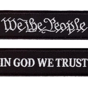 Op de afbeelding: Twee zwarte rechthoekige patches met wit geborduurde tekst. De bovenste patch leest "We the People" in een decoratief lettertype. De onderste patch leest "In God We Trust" in een schreefloos lettertype.