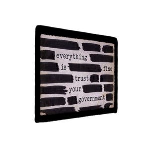 May include: A rectangular patch with a black border featuring a white background covered in horizontal black brushstrokes. The text "everything is fine trust your government" is printed in a black typewriter-style font across the white spaces.
