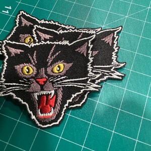 Op de afbeelding: Geborduurde patch met een zwarte kat met gele ogen, een rode mond en witte snorharen. De kat gromt, met zijn mond open. De patch heeft een witte rand en ligt op een groene ondergrond.