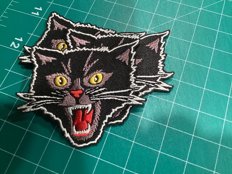 Op de afbeelding: Geborduurde patch met een zwarte kat met gele ogen, een rode mond en witte snorharen. De kat gromt, met zijn mond open. De patch heeft een witte rand en ligt op een groene ondergrond.