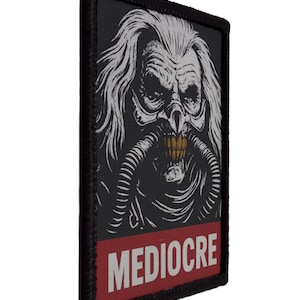 Op de afbeelding: Zwarte patch met een gedetailleerde illustratie van een grotesk gezicht met lang wit haar en een ademhalingsapparaat. Het woord "MEDIOCRE" is in het wit gedrukt op een rode banner aan de onderkant. De patch heeft een zwarte rand.