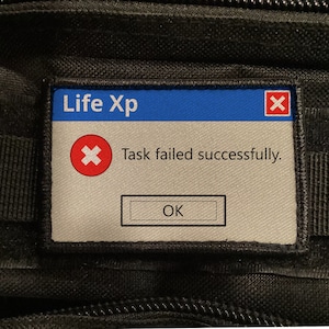Peut inclure: Un patch rectangulaire avec une section supérieure bleue indiquant "Life Xp" et un "X" rouge dans le coin supérieur droit. Le texte "Task failed successfully." est en dessous. Le patch a un bouton "OK".